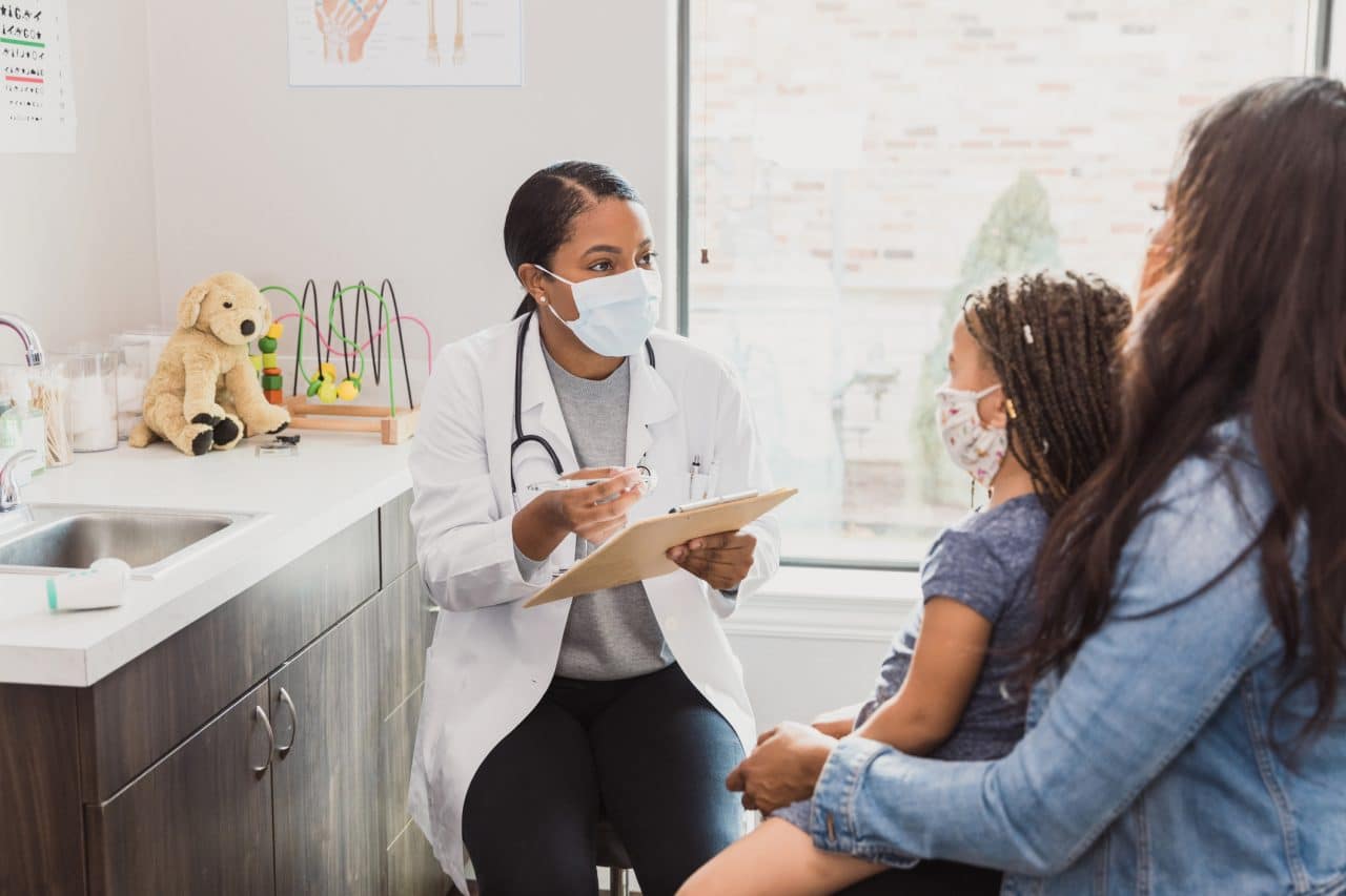 With a protective mask on, a female pediatrician talks to a young patient's mother about the woman's daughter's medical conditions. They are wearing protective masks during the COVID-19 pandemic.