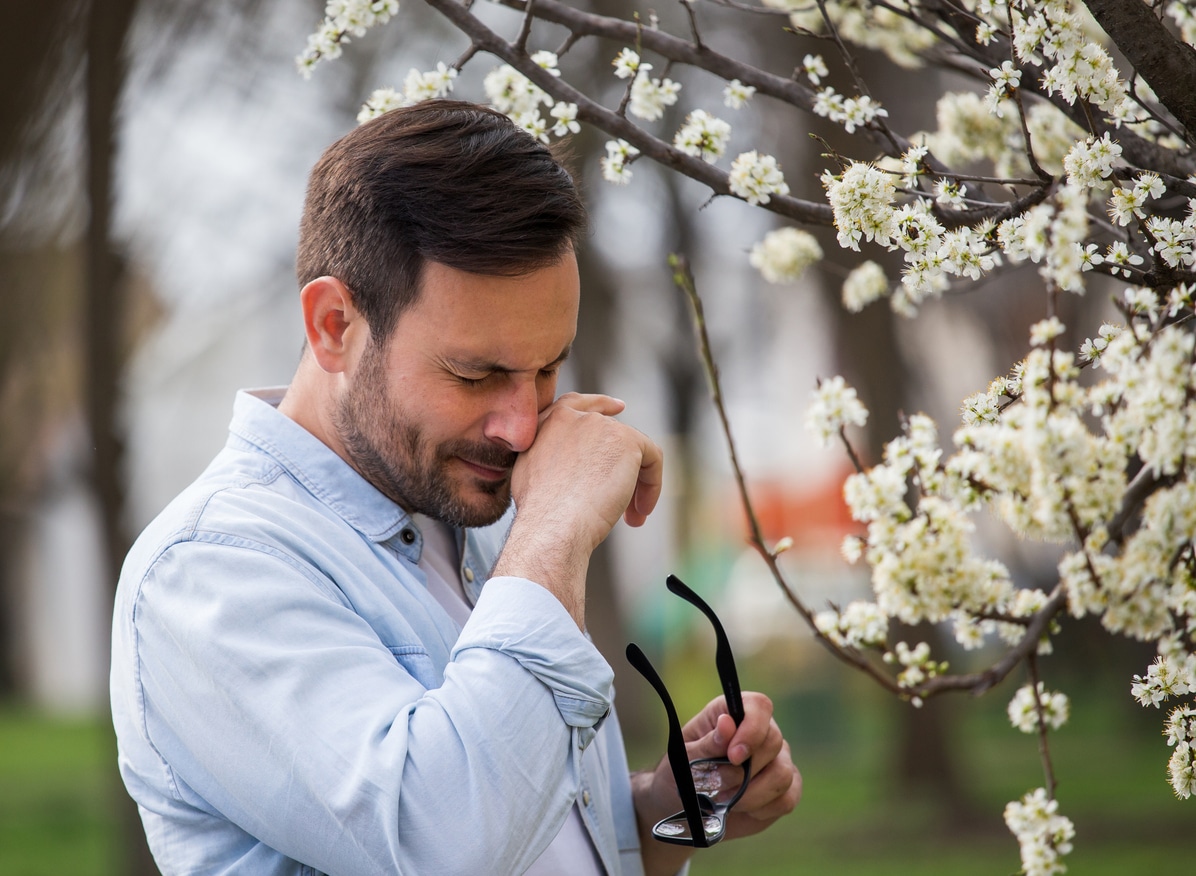 Man with pollen allergies rubbing his nose next to a flower tree.